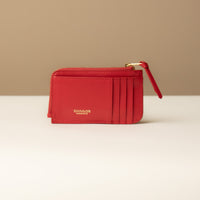 A red card case with a zipper coin compartment, made of polished pebble leather, with the letter 'D' visible on the front.