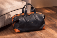 Black and brown duffel bag on a wooden floor next to a beige couch.