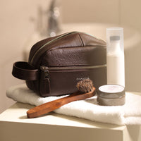 A brown toiletry bag 2014 with a silver zipper closure and a handle, with visible brand Danilos, placed in a bathroom. 