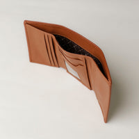 A open trifold wallet in tan color, made from pebbled leather, with nappa leather interior with slots for cards and a long comparment for cash, displayed against a neutral background. Upper view