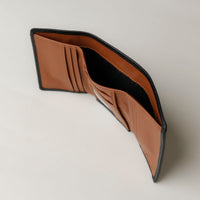 A open trifold wallet in black and tan color, made from pebbled leather with nappa leather interior, with 6 slots for cards and a long comparment for cash, displayed against a neutral background. Upper view. 