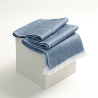 Folded blue blanket with white fringe on a light gray block against a white background