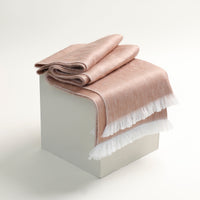 Folded beige blanket with white fringe on a white pedestal against a light gray background
