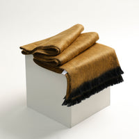 Folded brown blanket with black fringe on a white pedestal against a white background
