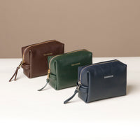 Three leather toiletry bags in brown, green, and blue colors, displayed against a neutral background.