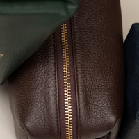 Close up image of Leather toiletry bags in brown, green, and navy colors, displayed against a neutral background.