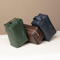 Three Leather toiletry bags in brown, green, and navy colors, displayed against a neutral background.