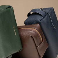 Close up view of three Leather toiletry bags in brown, green, and navy colors, displayed against a neutral background.
