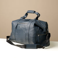 Angled view of A navy blue leather duffle bag with two handles and a zippered compartment on the front side.