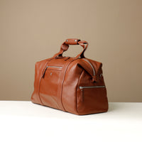 Angled View of a brown leather duffle bag with zippered compartment on the front side.