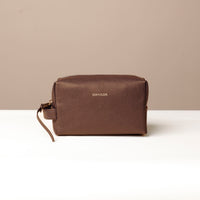 A brown, pebbled leather toiletry bag with a zipper closure and a brand logo embossed on the front. It is placed against a neutral background.