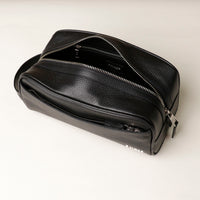 A pebble black, full-grain leather Emmiliano toiletry bag with a zipper closure, displayed against a neutral background. Open view. 