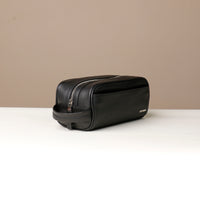 A pebble black, full-grain leather Emmiliano toiletry bag with a zipper closure, displayed against a neutral background. Lateral view. 
