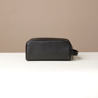 A pebble black, full-grain leather Emmiliano toiletry bag with a zipper closure, displayed against a neutral background. Back view. 