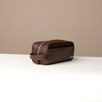 A pebble brown, full-grain leather toiletry bag Emmiliano with a zipper closure, displayed against a neutral background. Lateral view. 