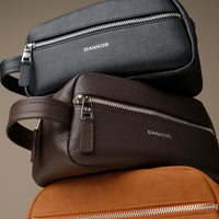 Three leather toiletry bags with zippered compartments, in black, brown, and tan colors, stacked on top of each other. With visible brand Danilos. 
