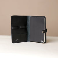 Front view of an open black Danilos pebble leather inskspirator with a clip closure, a notebook inside, and organized pouches for pens on a beige background