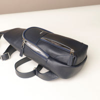 Close-up view of an open Danilos leather sling pack, Andres in a marine color with silver zippers on a beige background