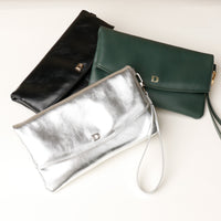 Front view of three leather Karelia Clutch Danilos in green, silver and black with a visible hand strap and gold accents over a white and beige background