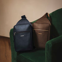 Two leather sling pack Andres, one black and one brown, on a green chair with a neutral background.