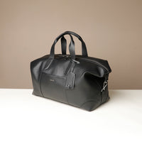 Side view of a black danilos hegarty travel bag with silver accents and tag over a beige background