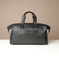 Back view of a black danilos hegarty travel bag with silver accents and a strap to attach to a suitcase, over a beige background