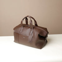 Side view of a brown danilos hegarty travel bag with silver accents and tag over a beige background