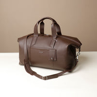 Side view of a brown danilos hegarty travel bag with silver accents, a travel tag  and a visible strapp, over a beige background