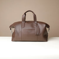 Front view of a brown danilos hegarty travel bag with silver accents and tag over a beige background