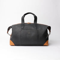A black leather duffle bag with two outside pockets and an adjustable leather strap, displayed against a neutral background.